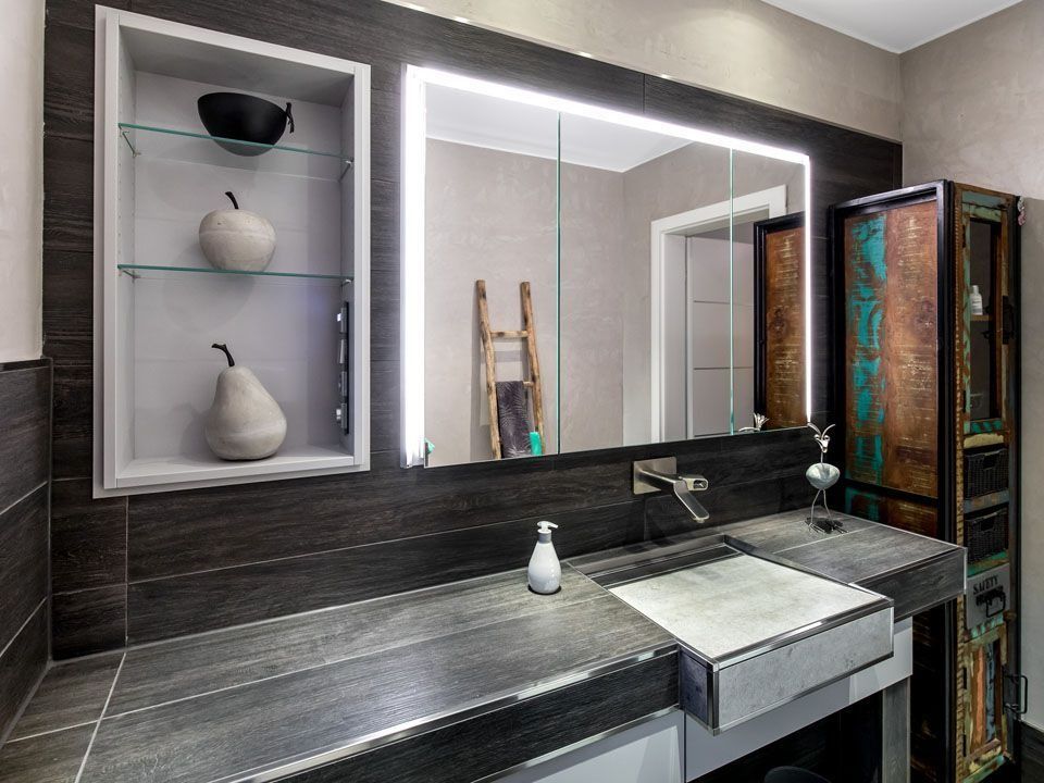 Modernes Badezimmer mit dunkler Arbeitsplatte, großem beleuchtetem Spiegel, eingebauten Regalen und antikem Schrank.