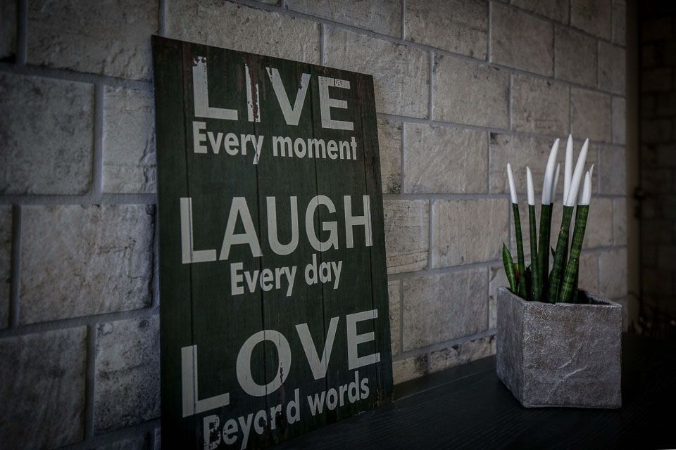 Ein Holzschild mit der Aufschrift „Lebe, lache, liebe“ an einer Steinmauer neben einer Topfpflanze.