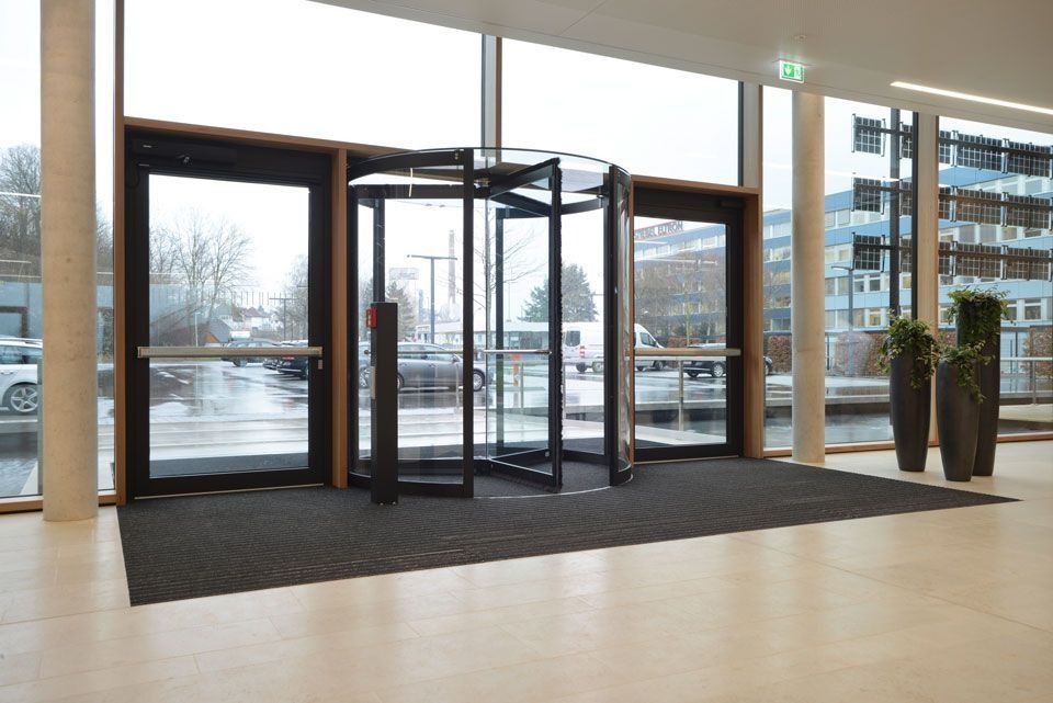 Drehtür im Eingang eines modernen Gebäudes mit großen Fenstern, die auf eine nasse Straße blicken.