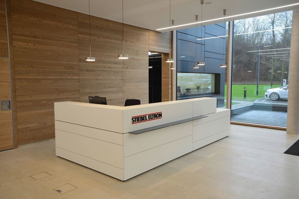 Moderner weißer Empfangstresen mit Firmenlogo, Akzentwand aus Holz und großem Fenster.