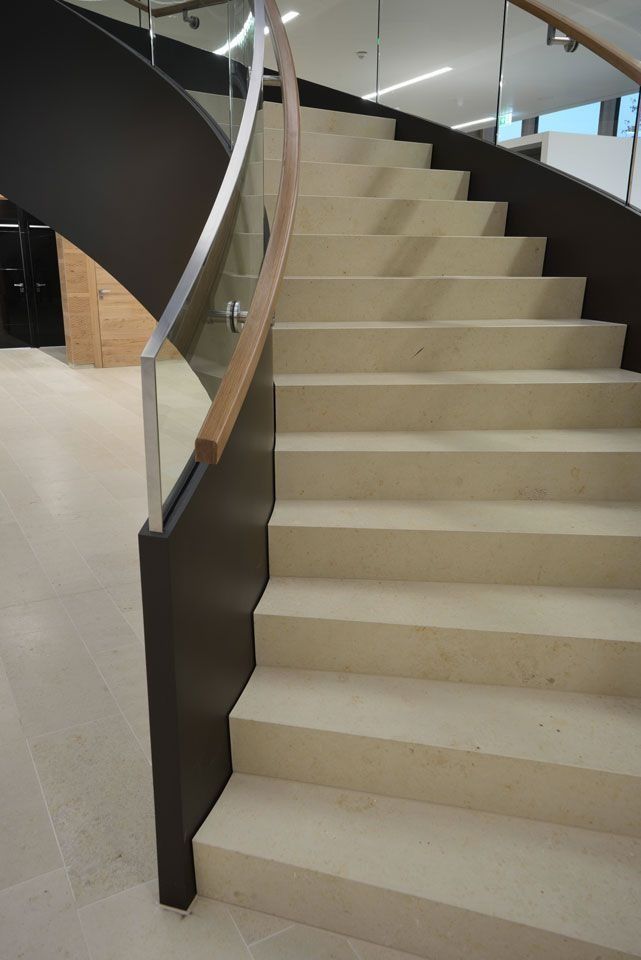 Geschwungene Treppe mit hellen Steinstufen, dunkelbrauner Seitenwand und Holzgeländer vor einer Glasbarriere.