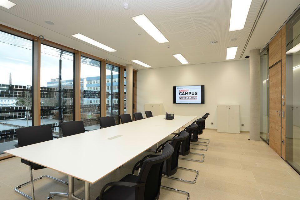Konferenzraum mit langem weißen Tisch, schwarzen Stühlen, großen Fenstern und einem an der Wand montierten Display.