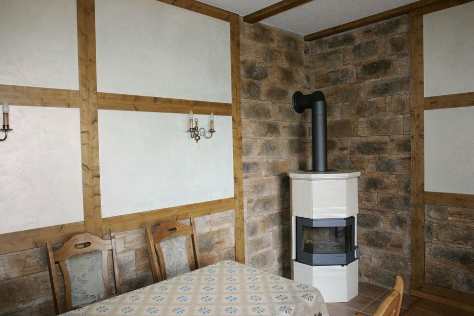 Ein Esszimmer mit Steinwand und weißem Ofen. Holzbalken rahmen weiße Putzwände ein.