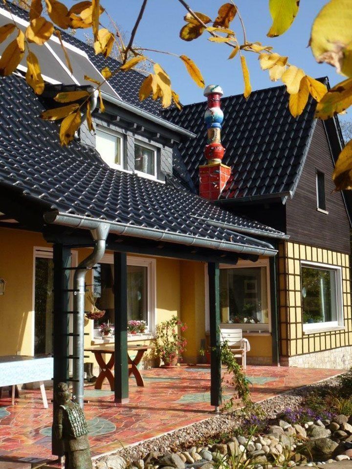 Gelbes Haus mit schwarzem Dach, überdachter Veranda und buntem Schornstein unter blauem Himmel.