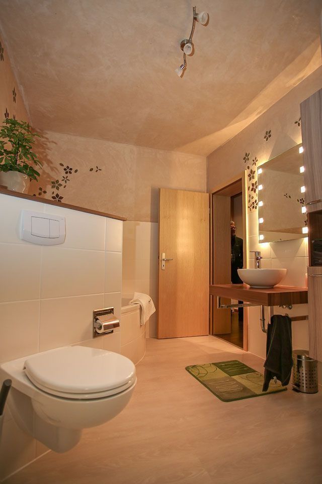 Modernes Badezimmer mit weißer Toilette, Waschbecken und Holztür. Beige Wände und Boden mit dekorativer Beleuchtung und Pflanzen.