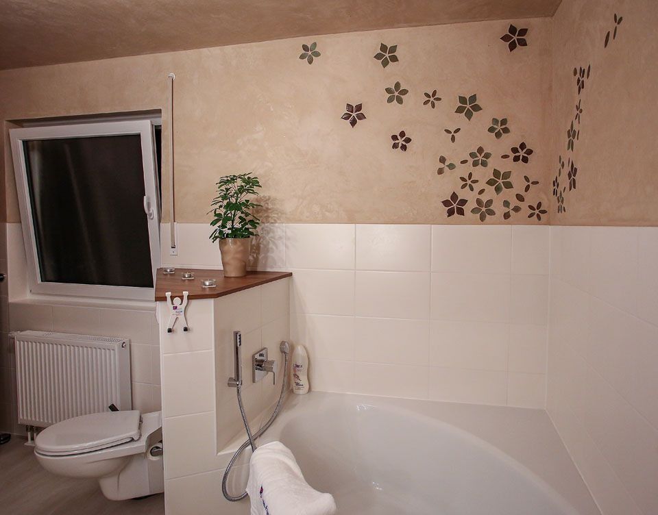 Badezimmer mit weißer Badewanne, Toilette und Fliesen. Beige Wand mit floralem Schablonenmuster. Fenster, Heizkörper und Pflanze.