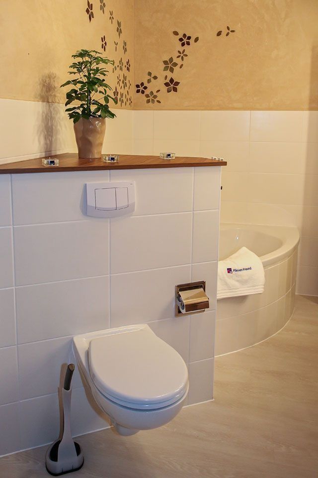 Badezimmer mit wandhängendem WC, weißen Fliesen und einem Holzregal mit einer Pflanze. Im Hintergrund ist eine Badewanne zu sehen.