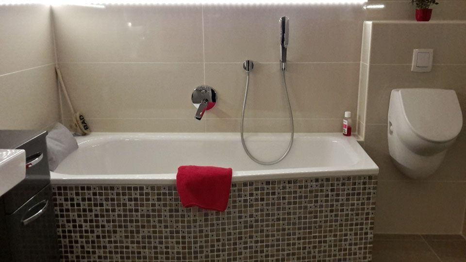 Badezimmer mit Badewanne, Duschkopf und Toilette, dekoriert mit Mosaikfliesen und rotem Handtuch.