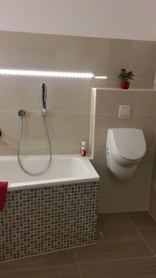 Badezimmer mit Badewanne, wandhängendem WC und beigefarbenen Fliesenwänden. Ein rotes Handtuch liegt auf der Wanne. Auf einem Regal steht eine kleine Pflanze.