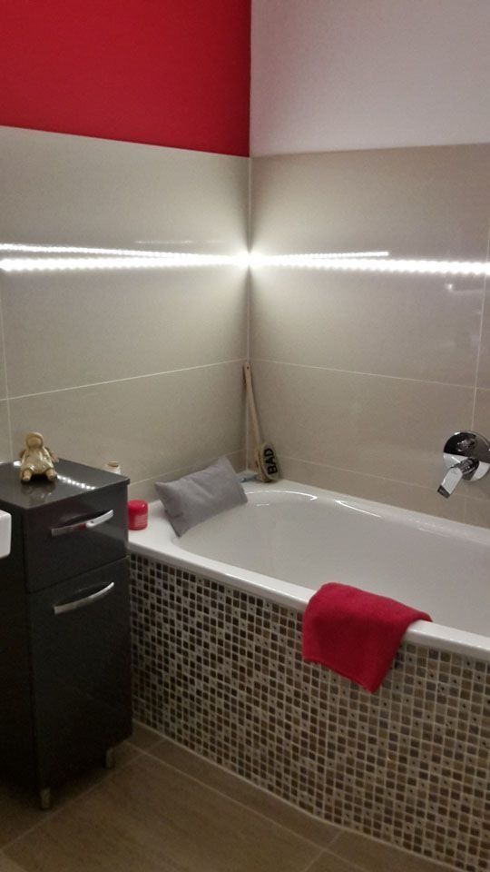 Badezimmer mit Badewanne, roter Akzentwand und Mosaikfliesen. LED-Streifenbeleuchtung und ein dunkler Schrank.