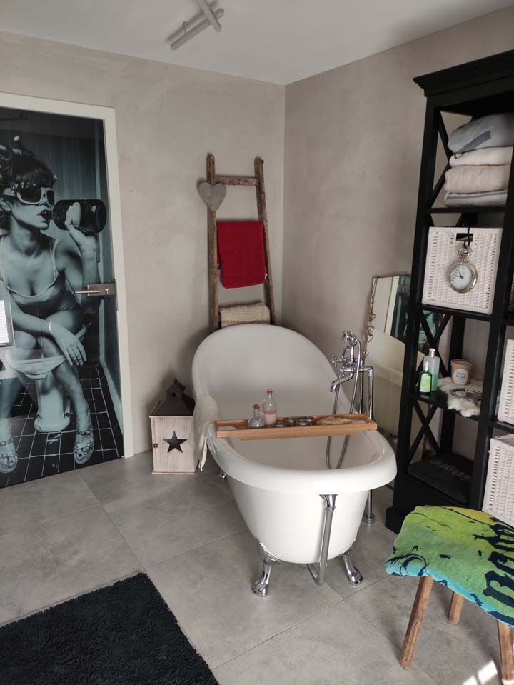 Badezimmer mit freistehender Badewanne auf Füßen, Handtuchleiter, offenen Regalen, einer Tür mit Kunstwerken und grauen Wänden.