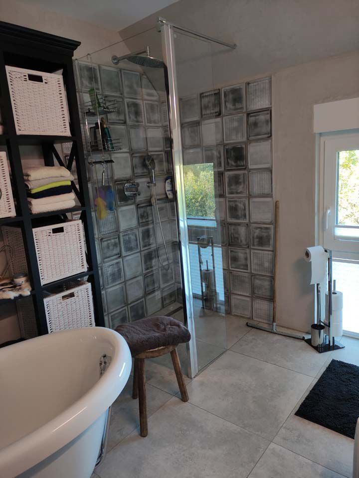 Badezimmer mit Glasdusche, gemusterter Wand, Ablageregalen und Badewanne.