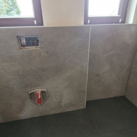 Grau geflieste Badezimmerwand mit flächenbündigen Armaturen und schwarzem Boden, neben einem Fenster.