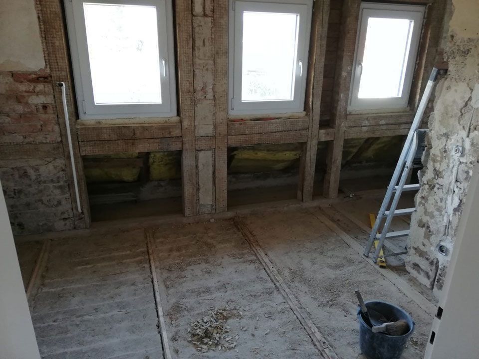 Innenraum im Bau, mit drei Fenstern, freiliegenden Holzbalken und einer Leiter.