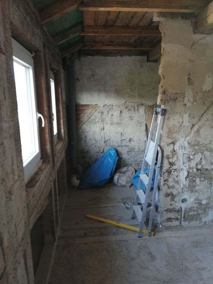 Innenraum im Bau, mit Fenstern auf der linken Seite und einer Leiter. Sichtbare, unverputzte Wände und Holzkonstruktion.