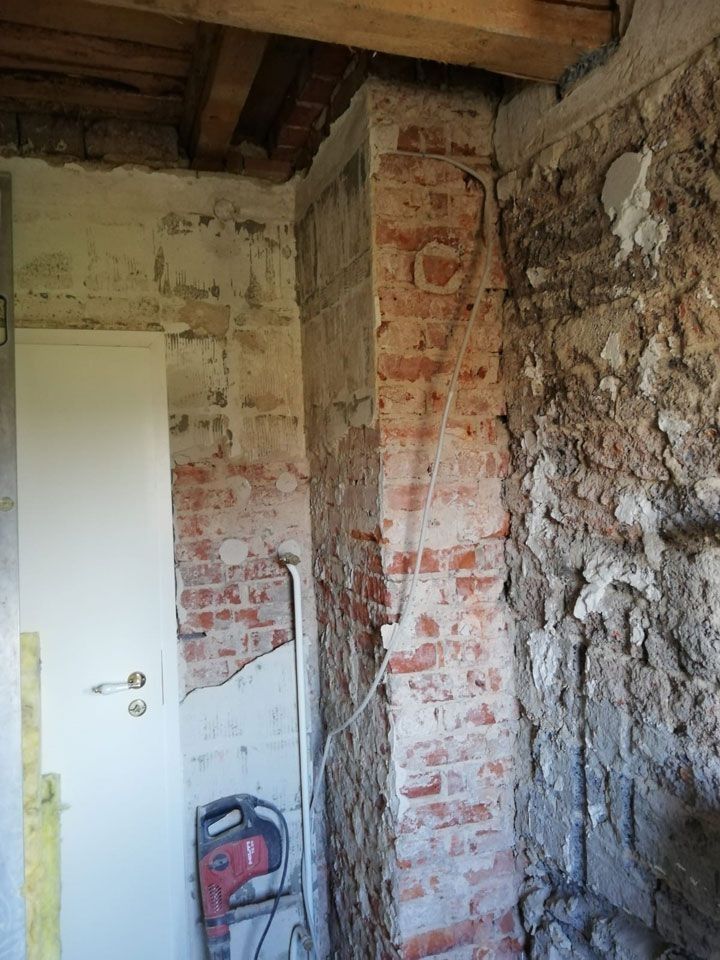 In einem im Bau befindlichen Raum sind eine gemauerte Säule und eine teilweise abgerissene Wand zu sehen, daneben eine weiße Tür und Werkzeuge.