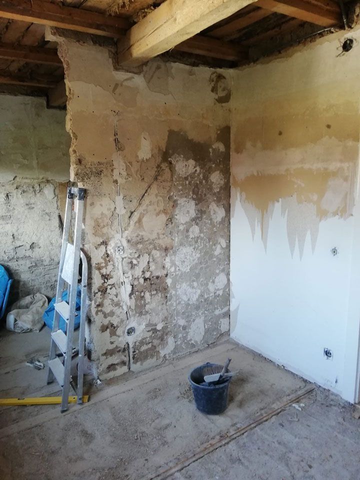 Innenansicht eines Zimmers, das gerade renoviert wird; Wände mit freiliegendem Putz, Leiter, Eimer und freiliegende Holzbalken.