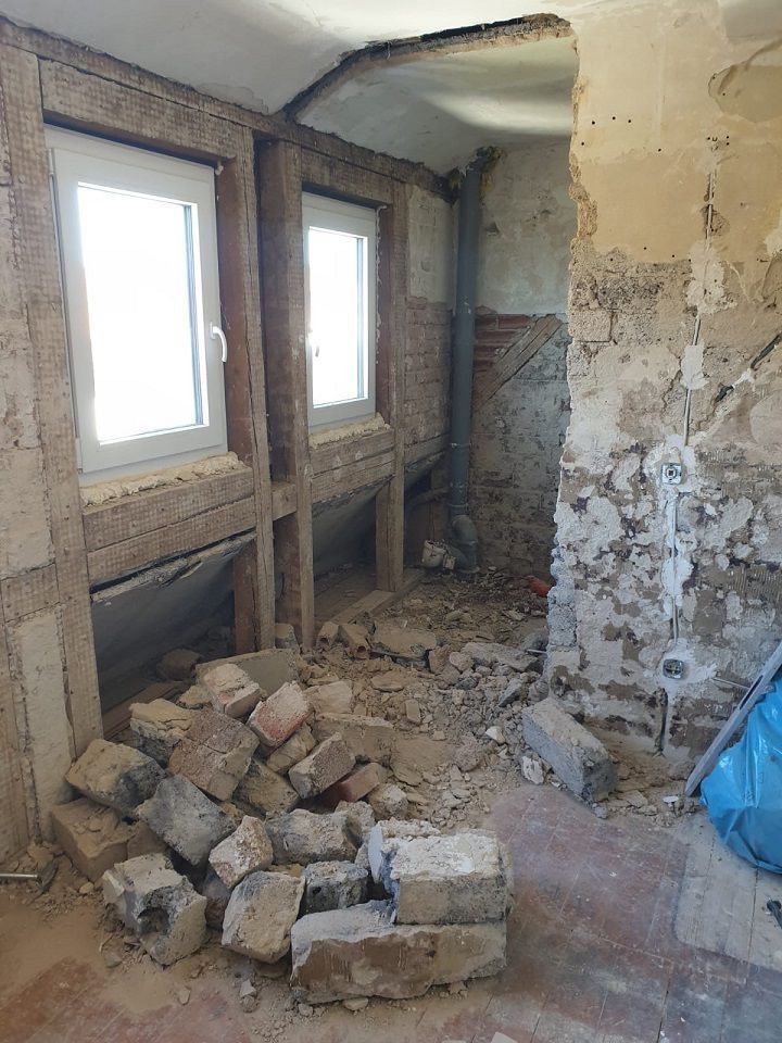 Zimmer im Umbau: Sichtbare Ziegelwände und Holzrahmen um die Fenster, Bauschutt auf dem Boden, unfertige Wände.