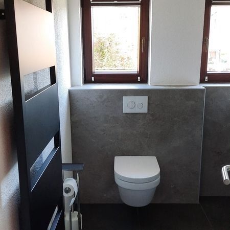 Modernes Badezimmer mit wandhängendem WC unterhalb der Fenster. Graue Wand, schwarzer Handtuchhalter links.