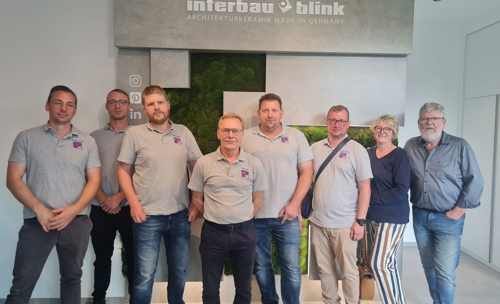 Eine Gruppe von neun Personen in Freizeitkleidung posiert vor einem Firmenschild vor einer grünen Wand.