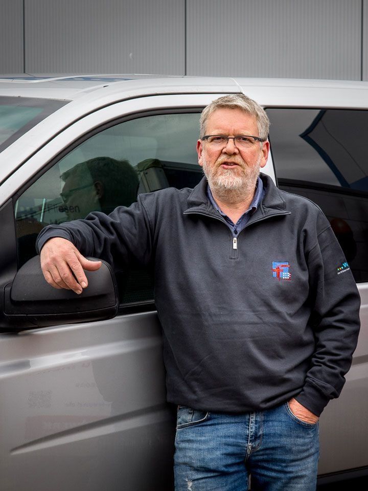 Mann mit Brille und dunklem Sweatshirt steht neben einem grauen Fahrzeug und lehnt an der Fahrertür.