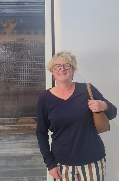 Eine Frau mit Brille lächelt und hält eine Handtasche. Sie trägt ein dunkelblaues Langarmshirt und eine gestreifte Hose. Sie lehnt an einer Wand mit Kunstwerken.