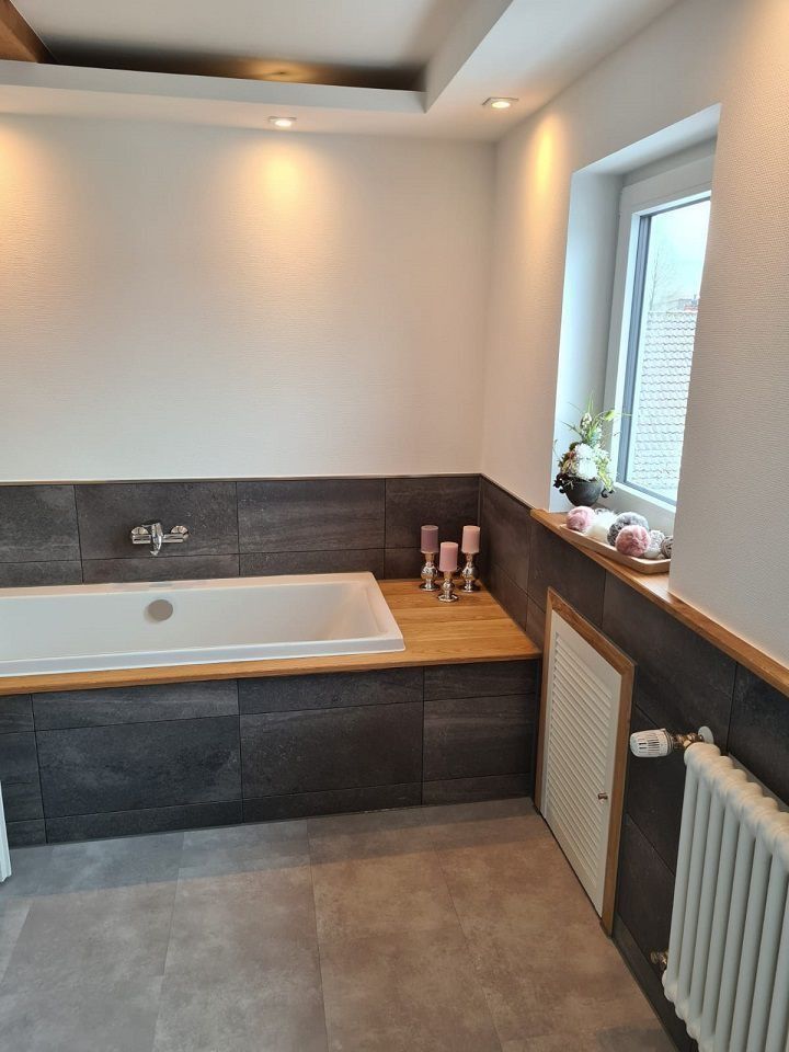 Badezimmer mit Badewanne, Holzelementen, grauen Fliesen und einem Fenster.