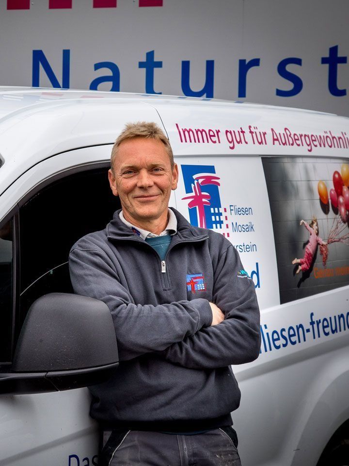 Ein Mann in Firmenuniform, die Arme verschränkt, lehnt an einem Firmenwagen mit der Aufschrift „Naturstein“ und „Fliesen-freunde“.