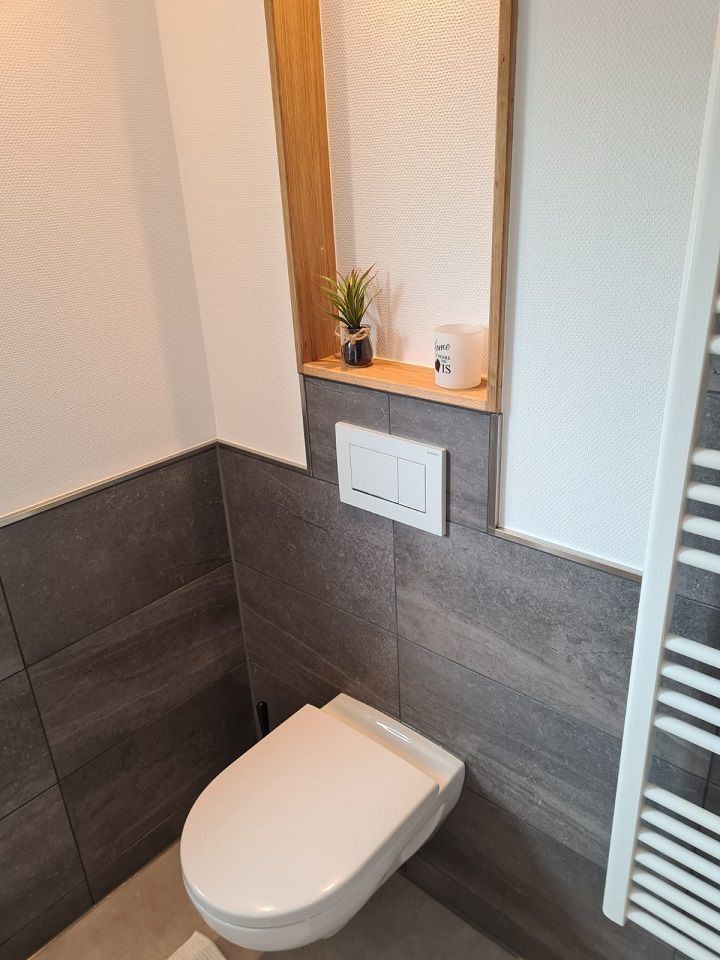 Toilette in einem modernen Badezimmer mit Holzregal, auf dem eine Pflanze und eine Kerze stehen. Grau geflieste Wände.