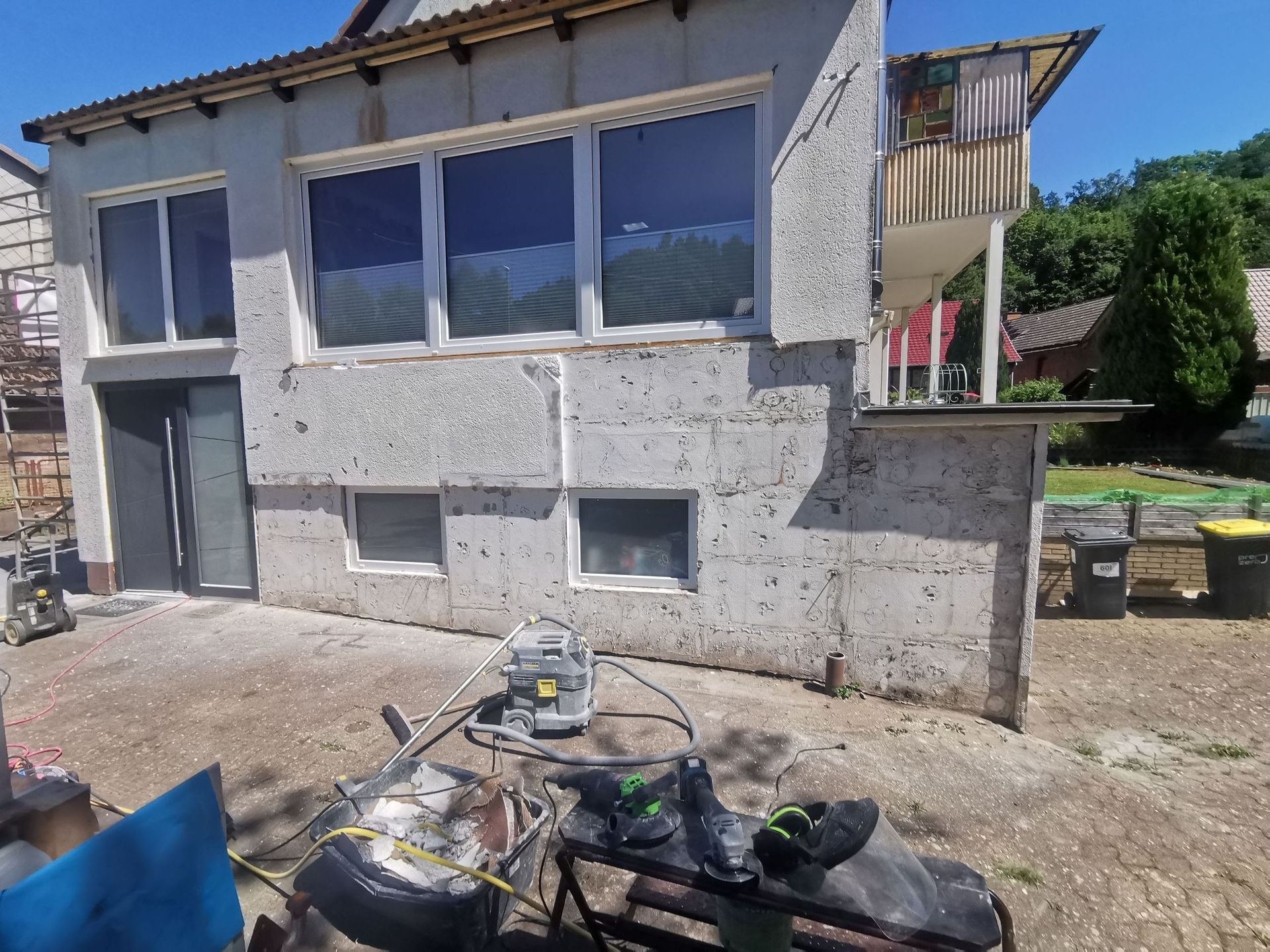 Zweistöckiges Haus mit unfertiger Außenisolierung; Werkzeuge und Bauschutt im Vordergrund; sonnige Außenkulisse.