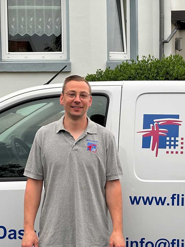 Ein Mann in grauem Hemd steht neben einem weißen Lieferwagen mit Logo und Website-Adresse.