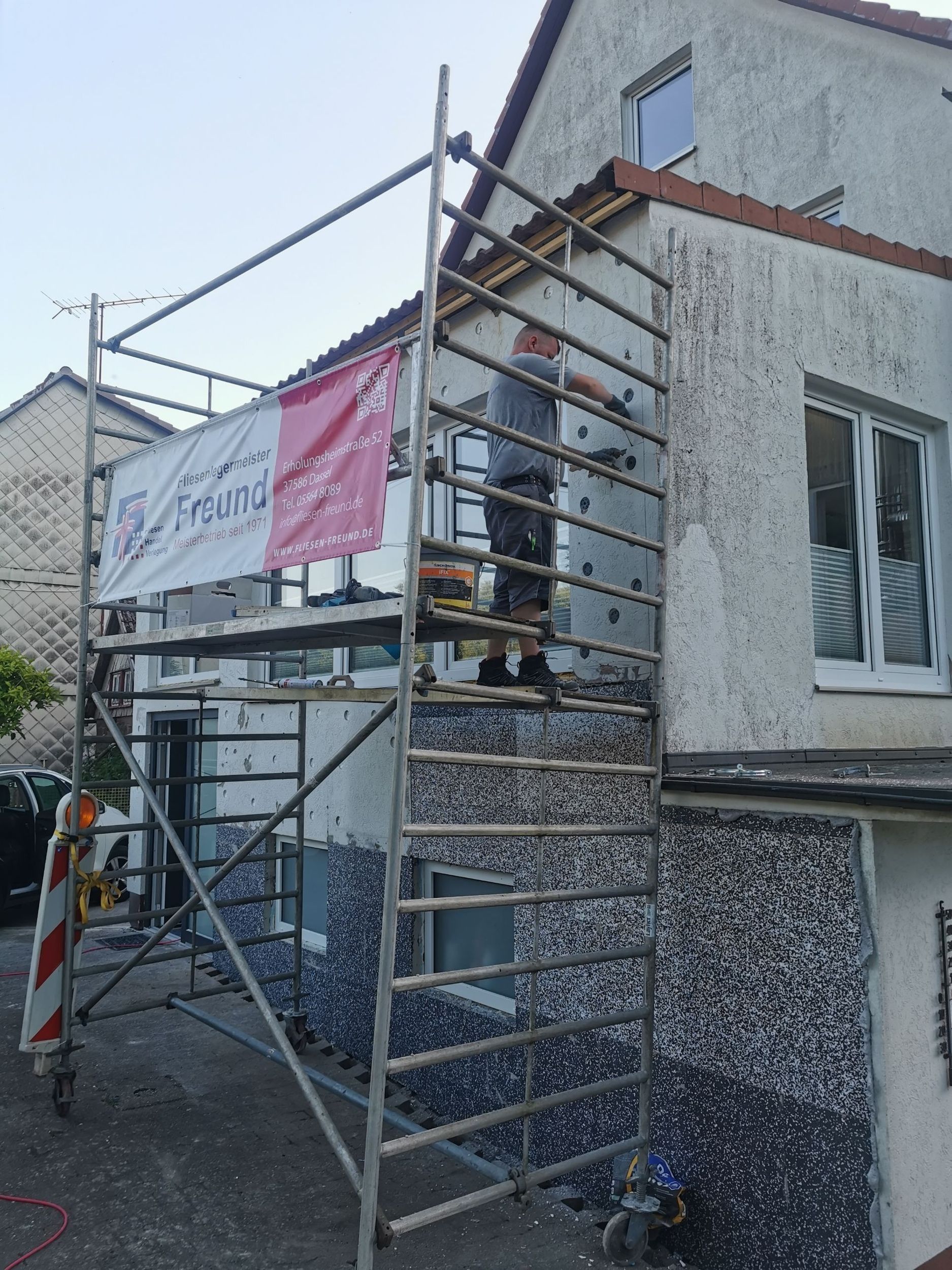 Mann auf einem Gerüst, der Dämmung an einer Hausfassade anbringt. Banner mit Logo sichtbar.