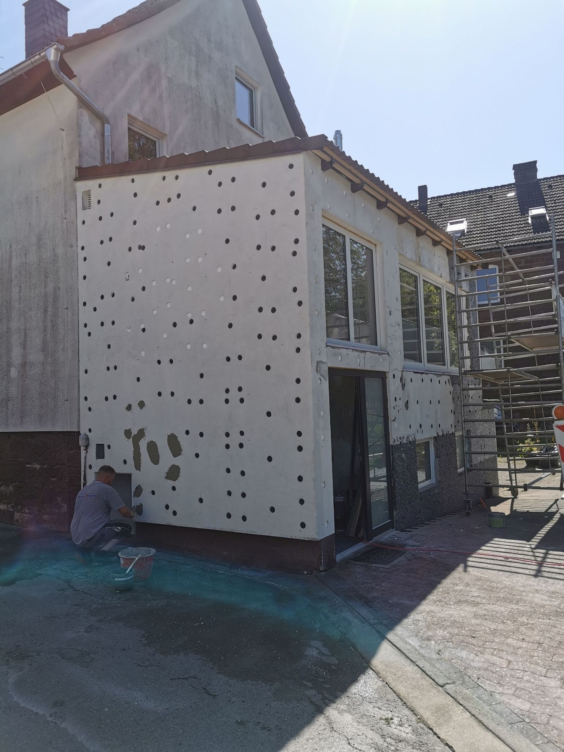 Außenansicht eines im Bau befindlichen Gebäudes; eine Person arbeitet an der mit Dämmmaterial bedeckten Wand, die viele Löcher aufweist.
