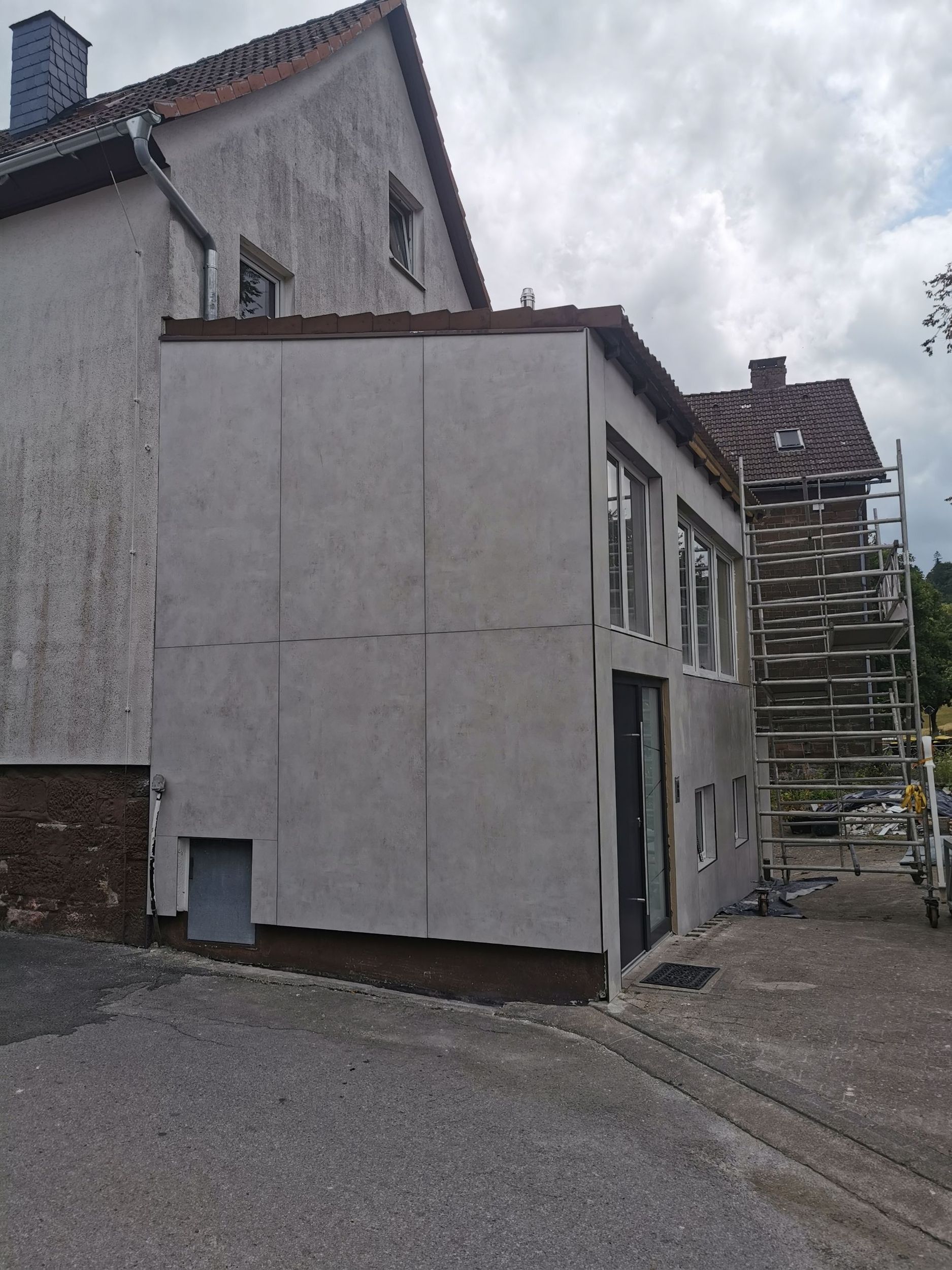 Außenansicht eines hellgrauen Gebäudes, das an ein zweistöckiges Gebäude angebaut ist. Rechts befindet sich ein Gerüst.