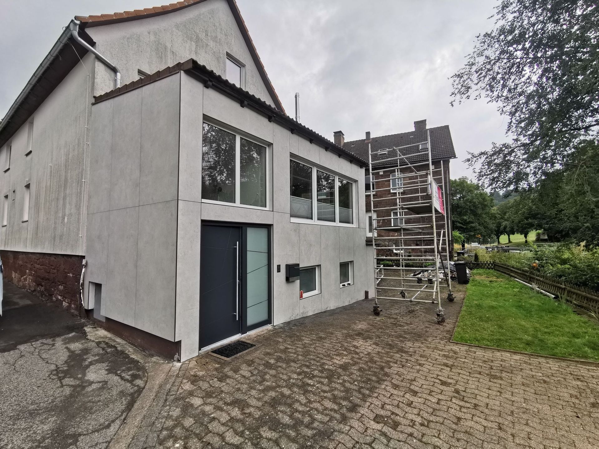 Graues Gebäude mit großen Fenstern und einer dunklen Tür, neben einem Gebäude mit Baugerüst.