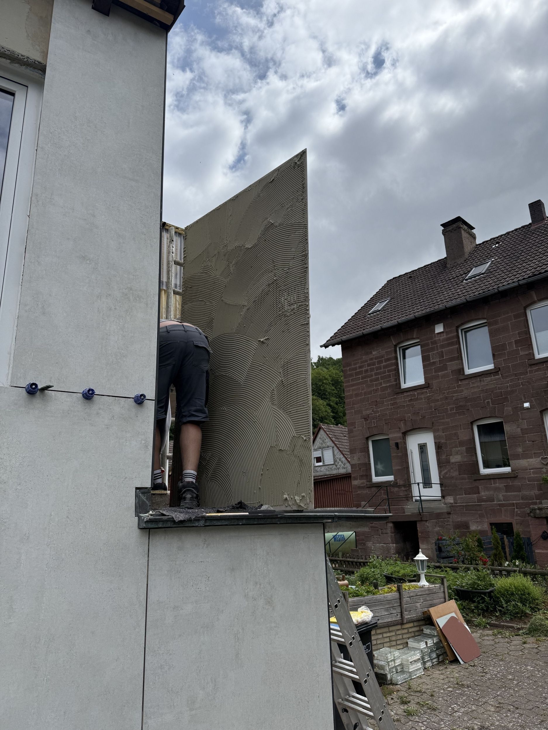 Person montiert an einem bewölkten Tag eine strukturierte Wandplatte an der Außenseite eines Gebäudes.