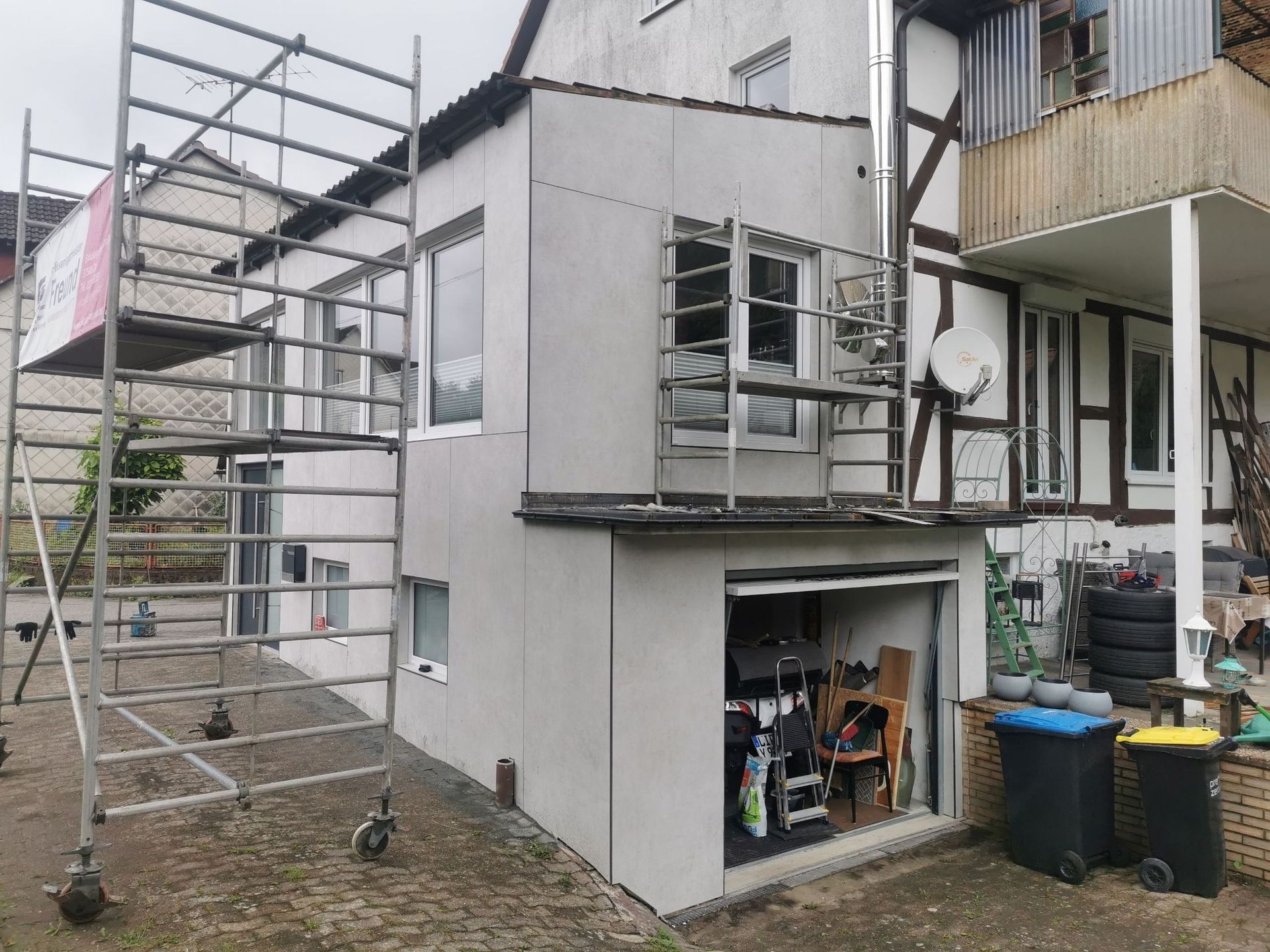Ein Baugerüst steht neben einem zweistöckigen Gebäudeanbau, das Garagentor ist geöffnet.
