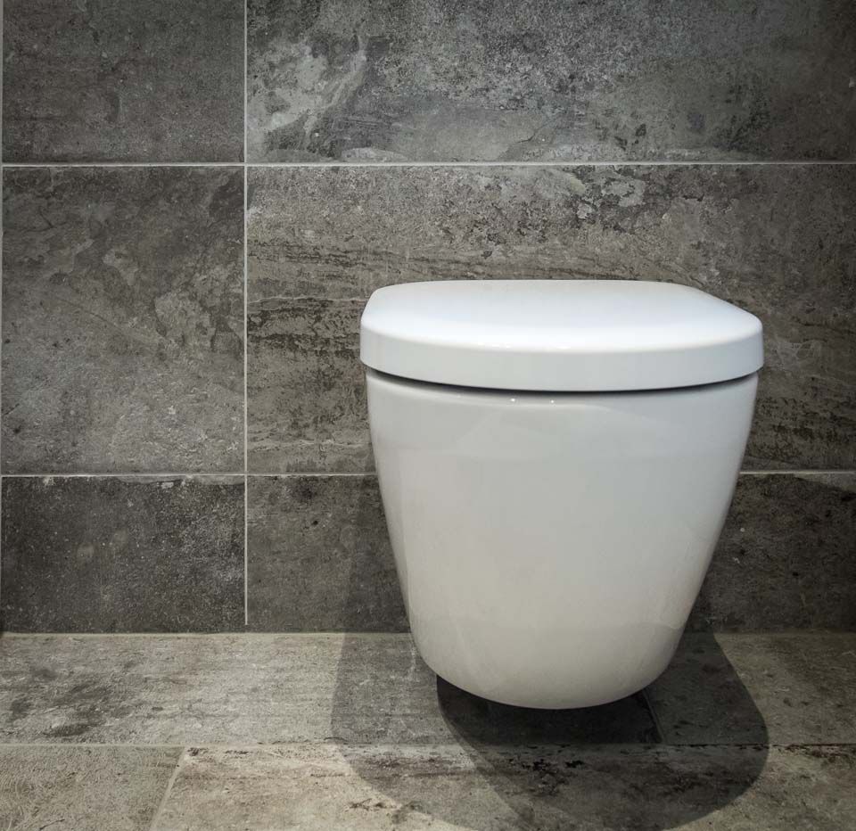 Weiße, wandhängende Toilette vor grauer Steinfliesenwand in einem Badezimmer.