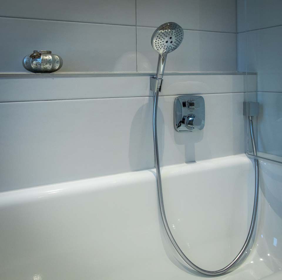 Weiße Badewanne mit Duschkopf und Bedienelementen, Ablage mit Dekorationsobjekt.