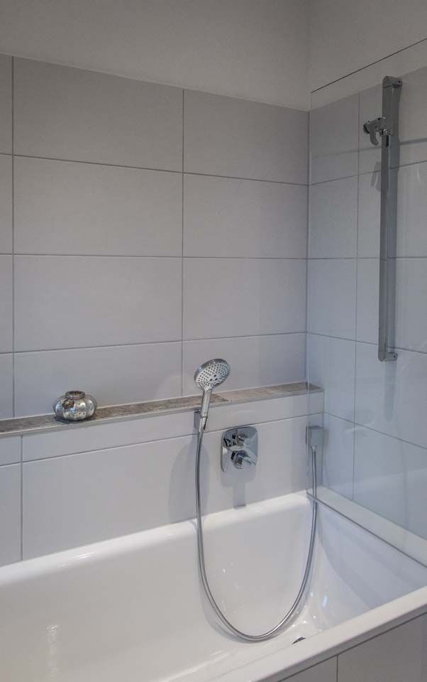 Weiß geflieste Dusche mit Badewanne, Duschkopf und Ablage.