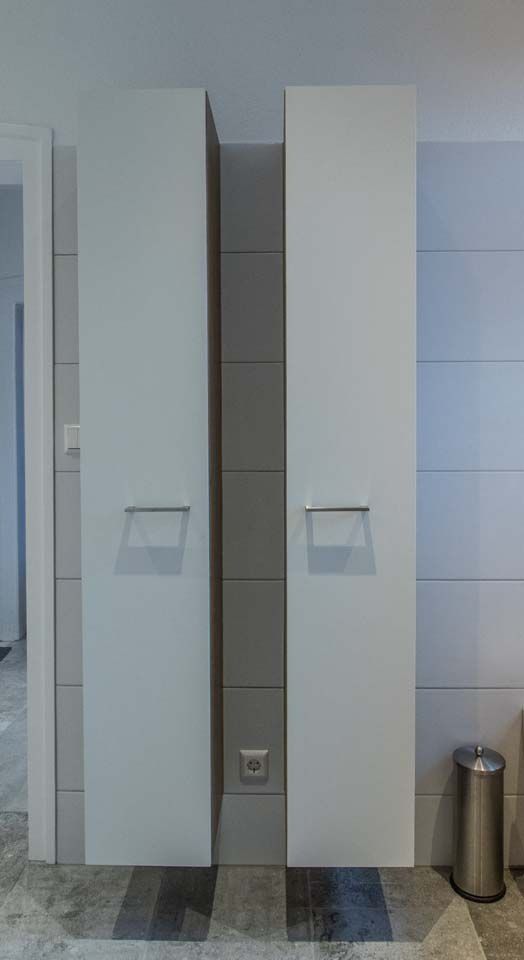 Zwei hohe, weiße Schränke flankieren einen grau gefliesten Wandabschnitt mit einer Steckdose.