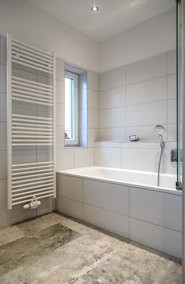 Weiß gefliestes Badezimmer mit Badewanne, Heizkörper und kleinem Fenster.