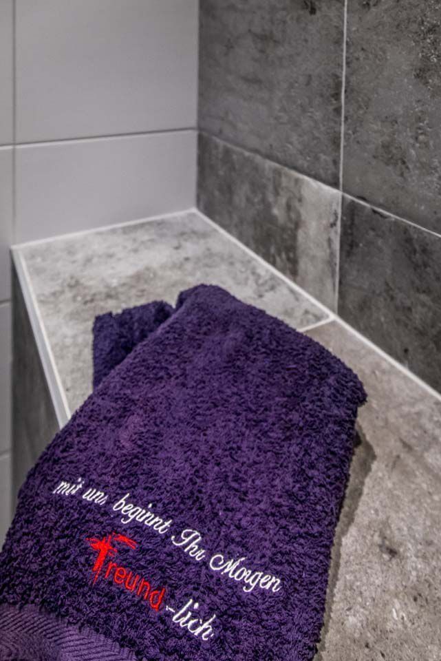 Ein violettes Handtuch mit der Aufschrift „Mit un beginnt der Morgen“ liegt auf einem gefliesten Regal im Badezimmer.