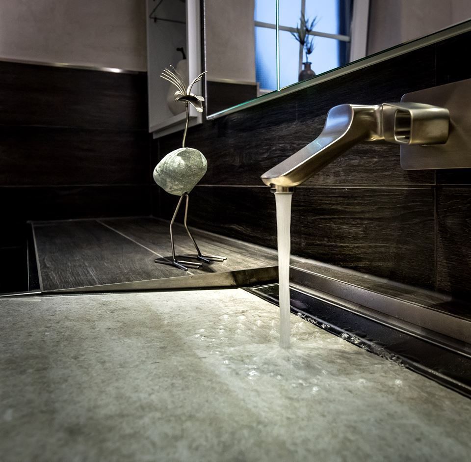 Modernes Badezimmerwaschbecken mit fließendem Wasser, skulpturalem Vogel, dunklen Fliesen und Fensterspiegelung.