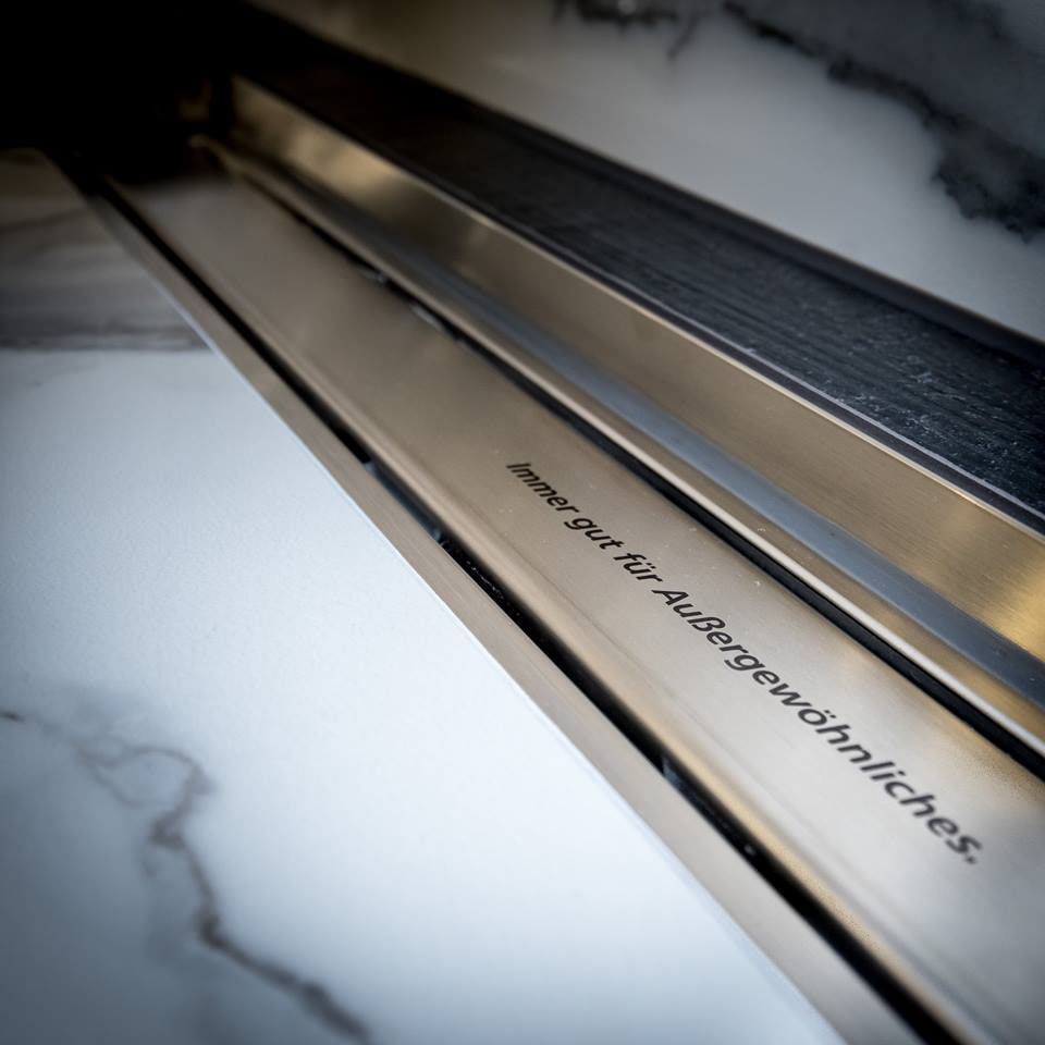 Nahaufnahme einer gebürsteten, linearen Duschrinne aus Metall mit eingraviertem deutschen Text. Marmorfliesen und ein dunkler Rost sind zu sehen.