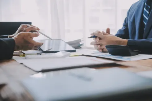 Dos personas de traje en un escritorio, revisando documentos y una tableta; discusión enfocada.