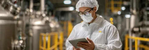 Persona con bata de laboratorio, gafas y mascarilla sosteniendo una tableta en un entorno de fábrica.
