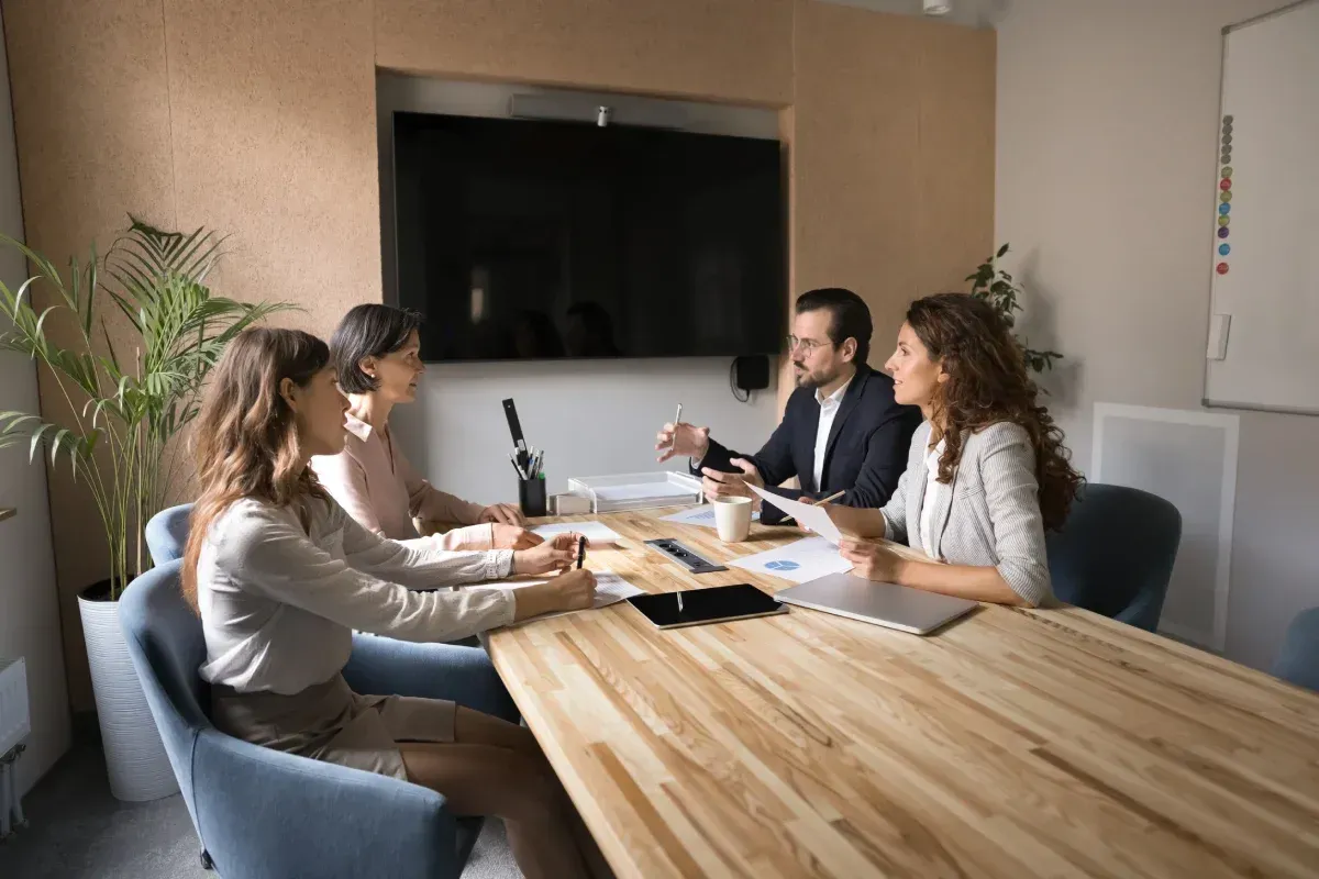 Cuatro personas en una reunión, alrededor de una mesa de madera. Un hombre gesticula mientras habla; los demás escuchan.
