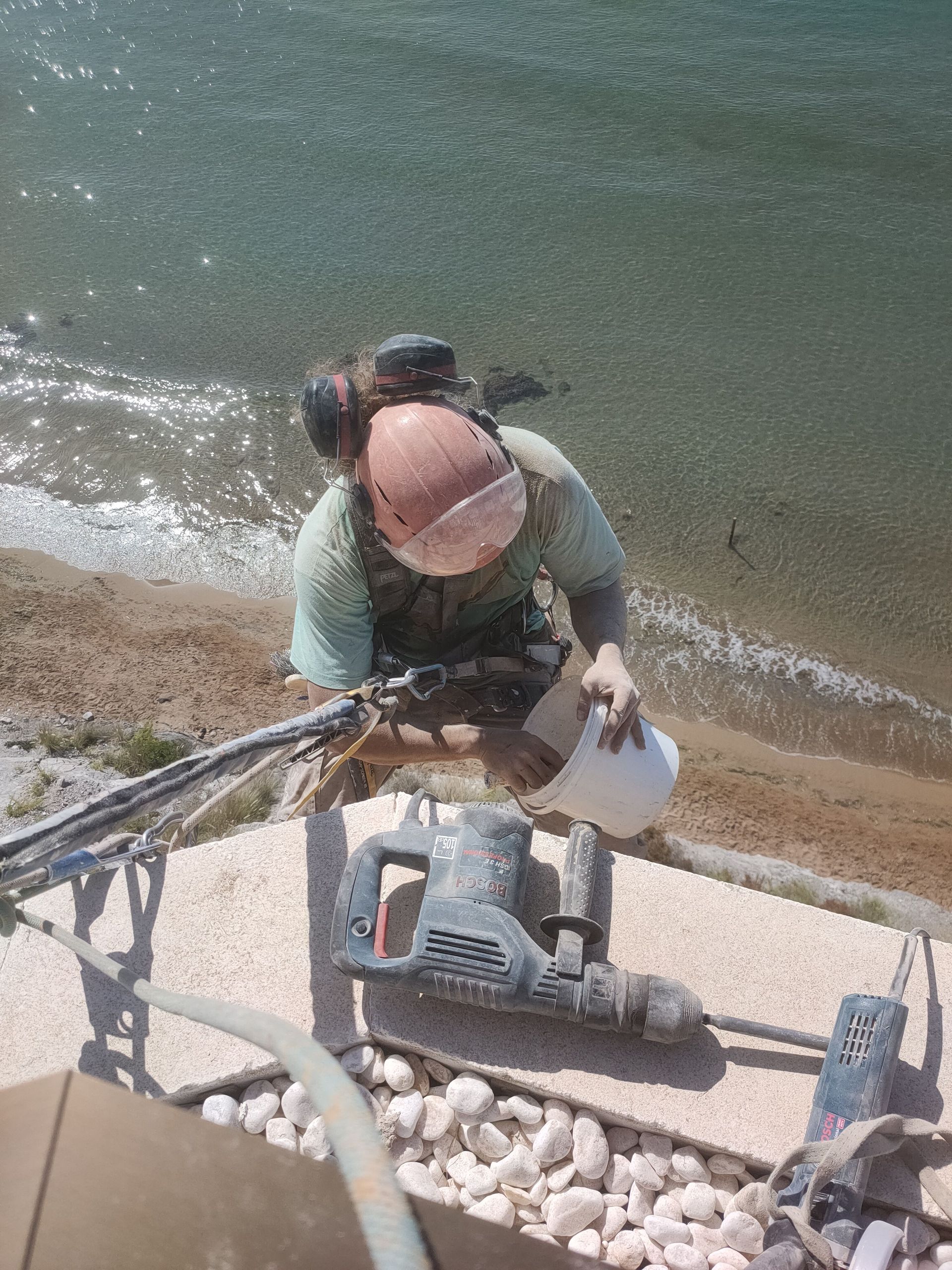 Hombre utilizando una herramienta eléctrica en el borde de un edificio cerca del agua. Lleva equipo de seguridad.
