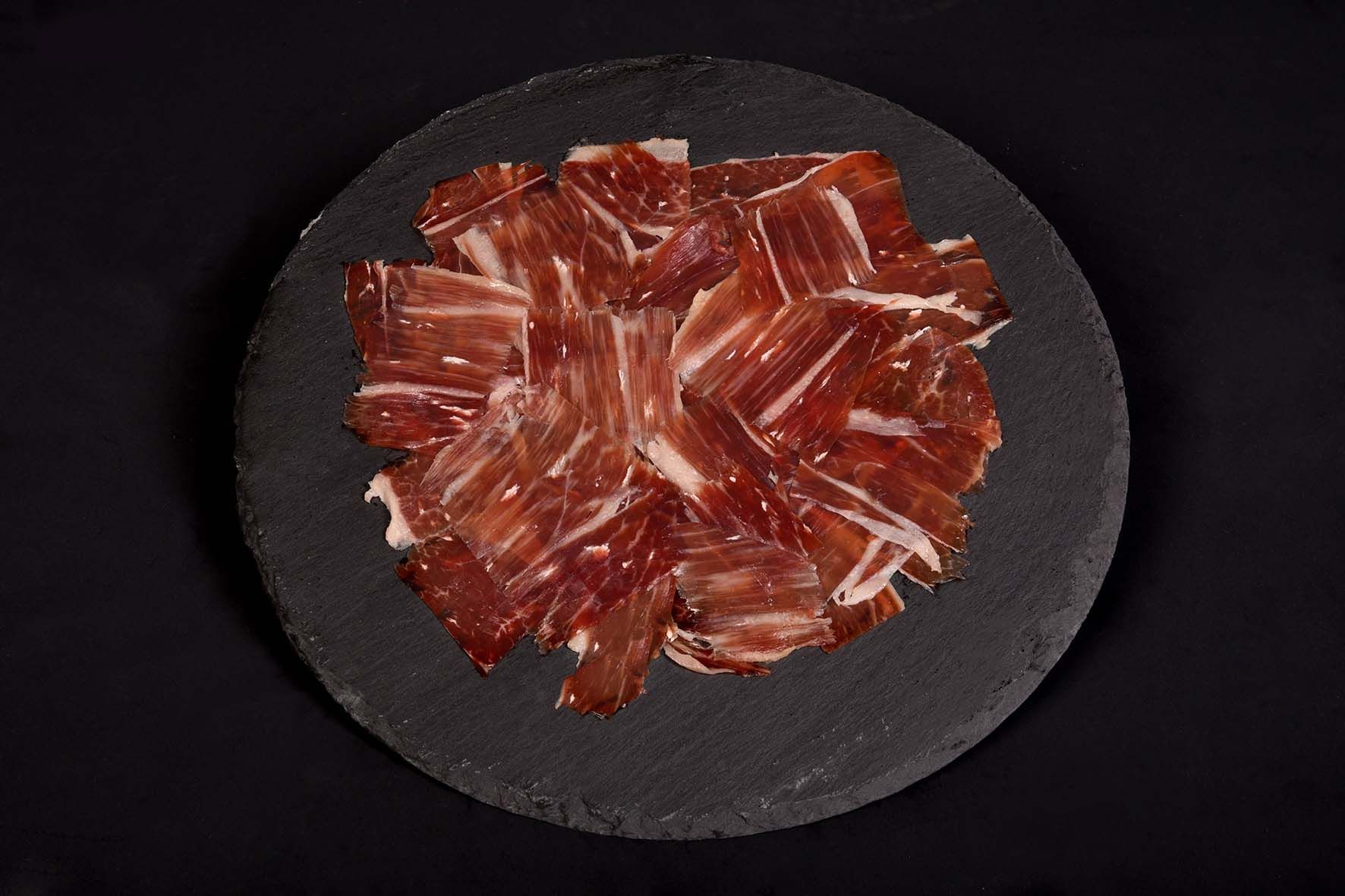 Jamón curado cortado fino sobre una bandeja redonda de piedra oscura, sobre un fondo negro.